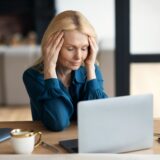Sad mature lady sitting at table in front of computer, suffering from headache, menopause, stress in